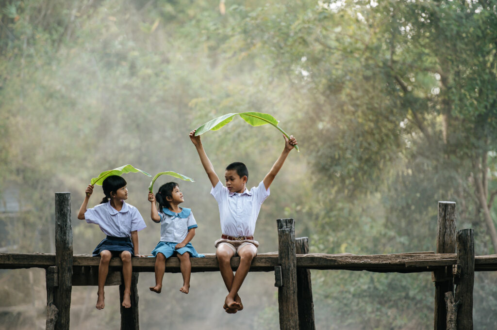 Three children sit barefoot on a rustic wooden bridge, joyfully playing with large green banana leaves under soft sunlight, surrounded by lush greenery—capturing a moment of innocent happiness and harmony with nature.