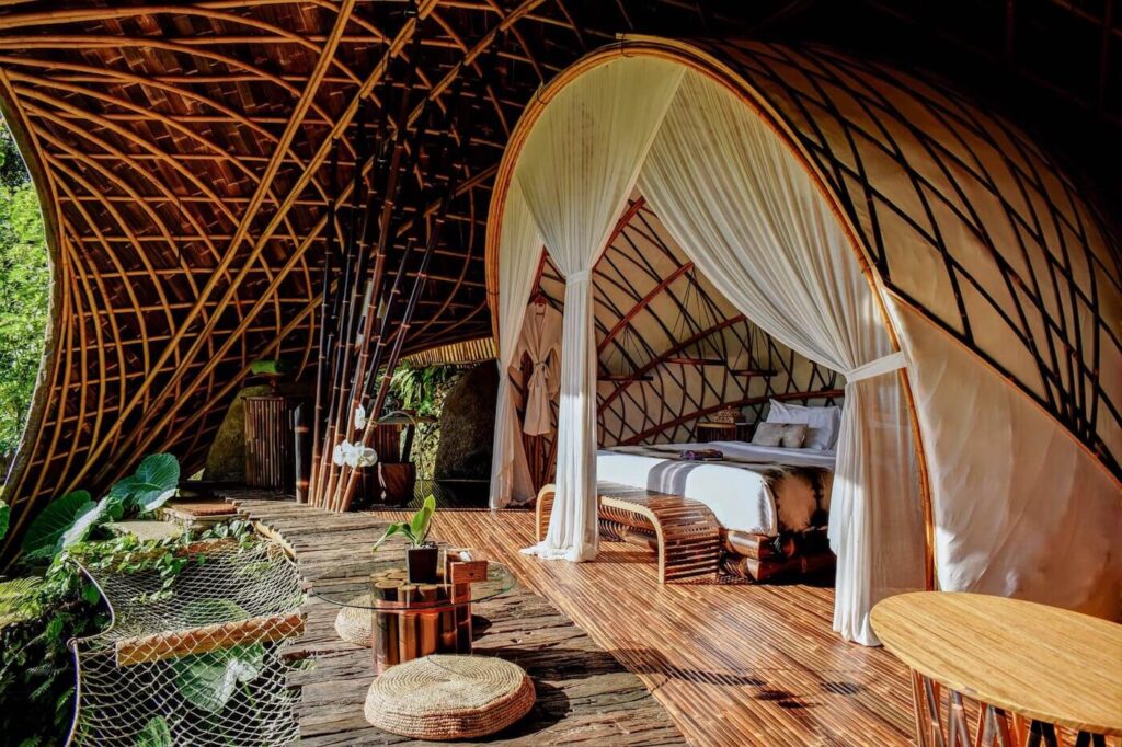 Sustainable bamboo villa in Bali offering a luxurious and eco-friendly retreat. A luxurious eco-friendly bamboo villa with an open-air design, featuring a canopy bed draped in white curtains, a wooden deck with woven seating, and lush greenery surrounding the space.