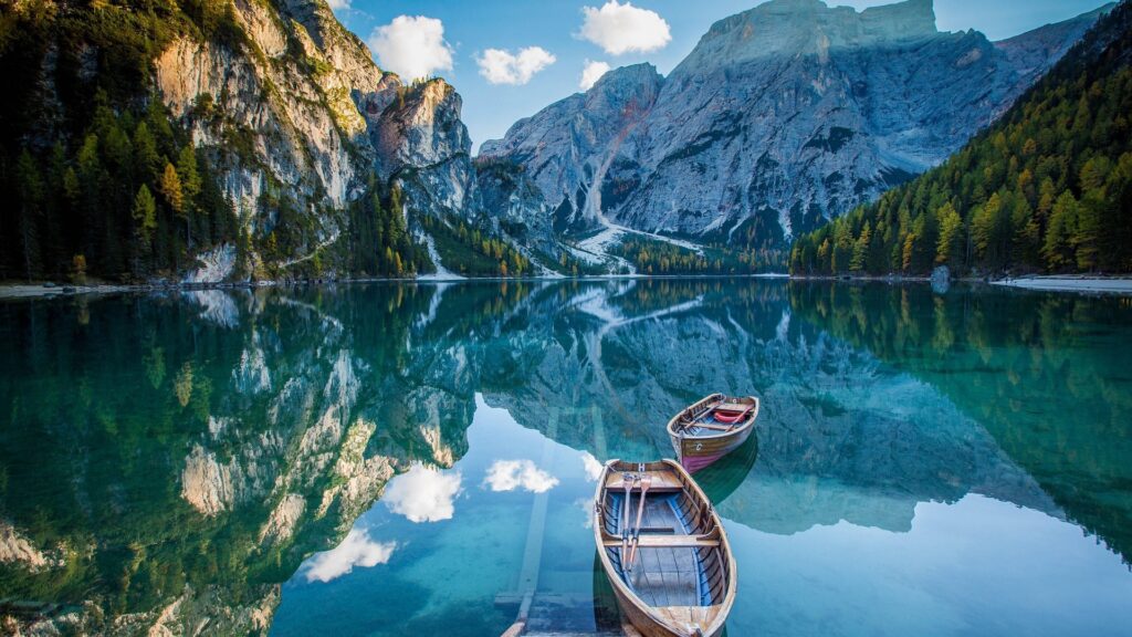 A breathtaking view of a serene alpine lake surrounded by towering mountains and lush green forests. Two wooden boats rest on the crystal-clear water, reflecting the beauty of the untouched natural landscape. This image embodies the essence of eco-friendly travelling, encouraging mindful exploration while preserving the planet’s pristine beauty.