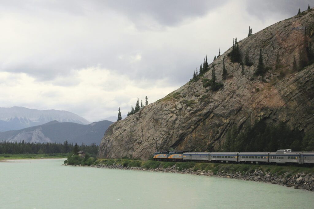 scenic train journey along a turquoise river, passing through rugged mountain landscapes under a cloudy sky. offering a lower carbon footprint while allowing passengers to enjoy breathtaking natural views.