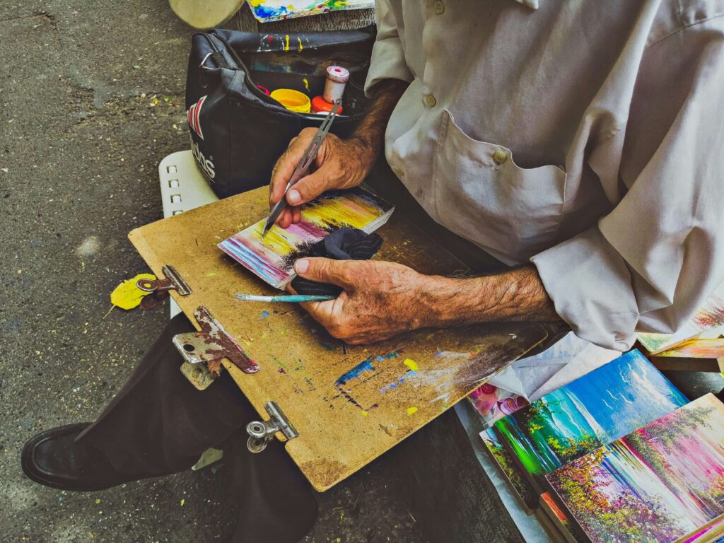 Skilled local artisan creating intricate handmade paintings with precision and passion An elderly artisan painting a small, colorful landscape artwork on a wooden clipboard, using fine brushes and a palette knife, surrounded by vibrant handmade paintings and art supplies