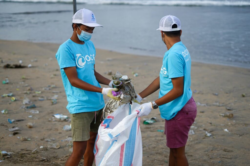 Two volunteers in blue shirts and gloves participate in a beach cleanup, collecting plastic waste and debris. Supporting local clean-up efforts is a crucial step toward eco-friendly travelling, ensuring destinations remain pristine for future generations.