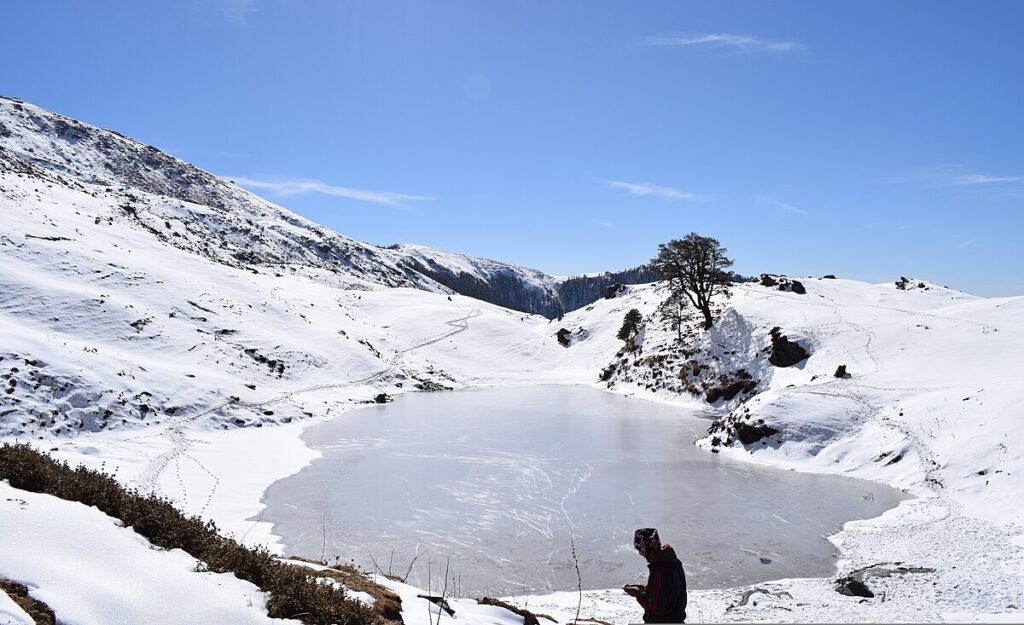 A tranquil winter scene with a snow-laden lake and lone figure, representing the pristine beauty of eco-friendly treks in the Indian Himalayas.