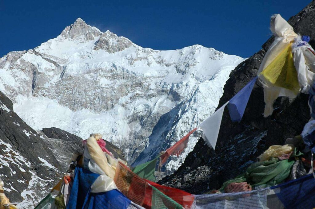 Colorful prayer flags flutter at Goecha La Pass with Mount Kanchenjunga towering in the background, symbolizing the spiritual and natural harmony of sustainable Himalayan trekking in Sikkim.