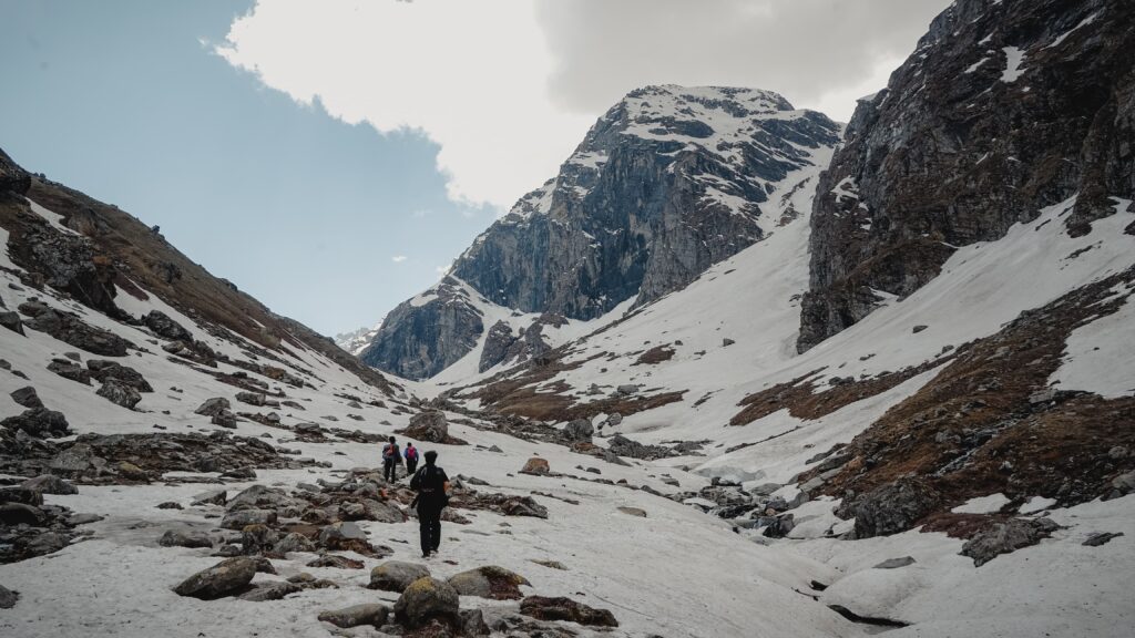 Trekkers walking through a snow-covered valley surrounded by steep rocky mountains near Hampta Pass, with patches of brown earth and a bright sky overhead, capturing the rugged beauty and adventure of the Himalayan landscape.