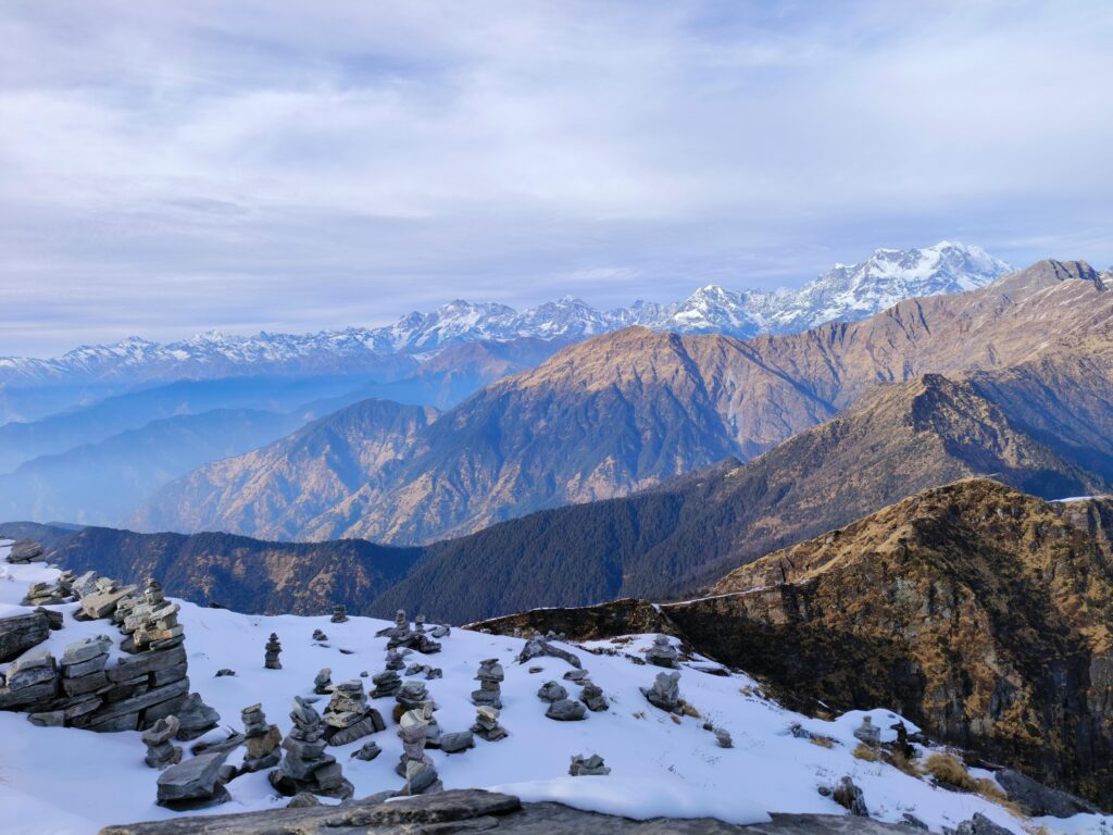 A panoramic view of the snow-dusted Himalayan range with stacked stone cairns in the foreground and layers of rugged, forested mountains fading into the distance—capturing the serene essence of the Top 10 Treks in India for Sustainable Travel, where nature meets mindful exploration.